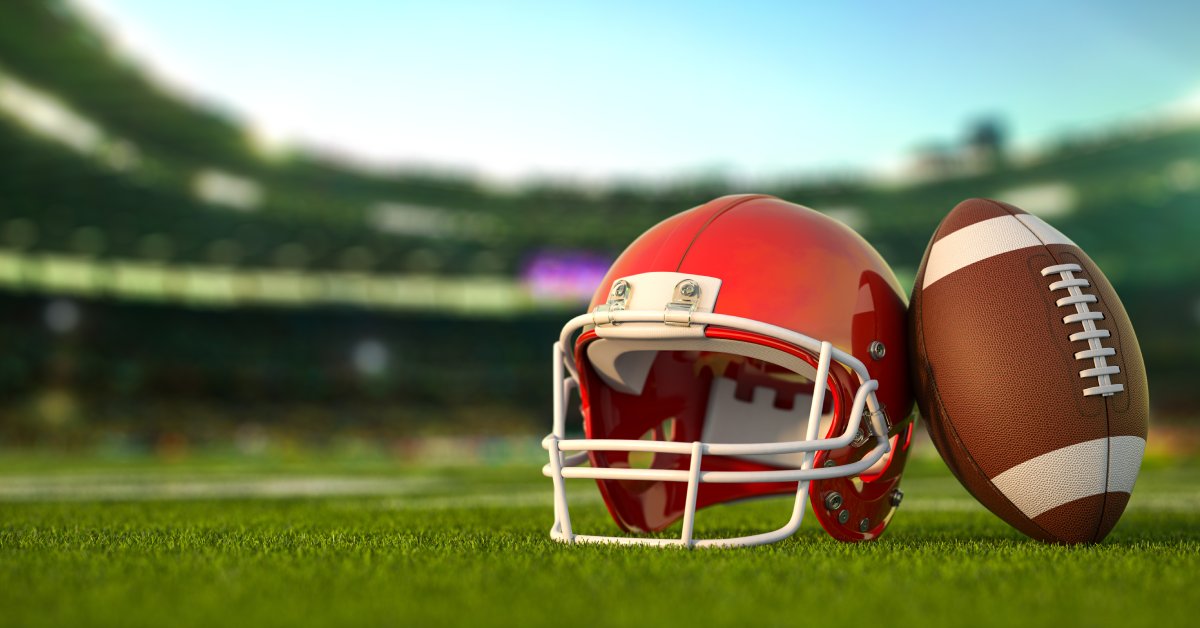 A red American football helmet with a white facemask sitting on grass field in a stadium with a football learning against it.