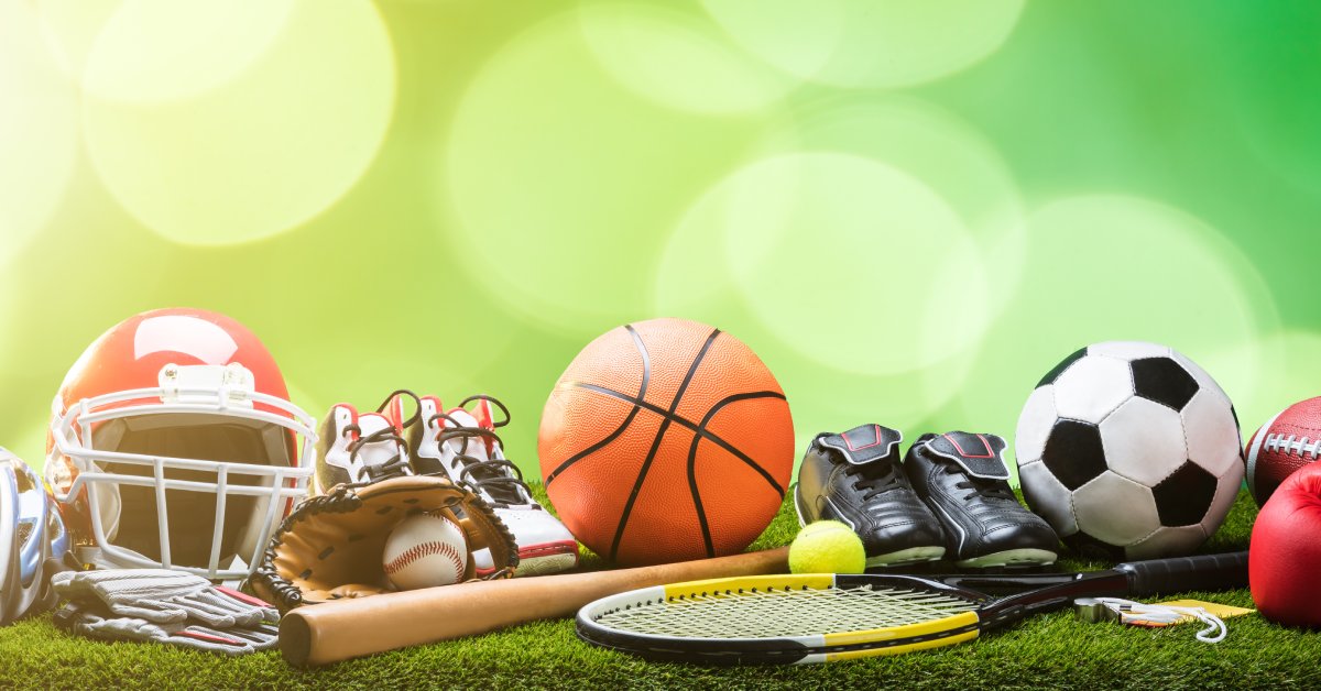 A set of various sports equipment, including a basketball, baseball and glove, tennis racquet, and more on a grass surface.