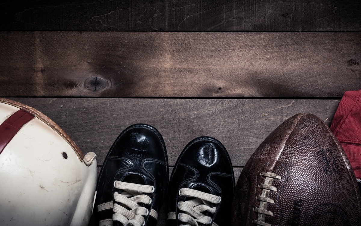An overhead view of old, vintage football gear including a white helmet, black cleats, leather football, and red jersey.