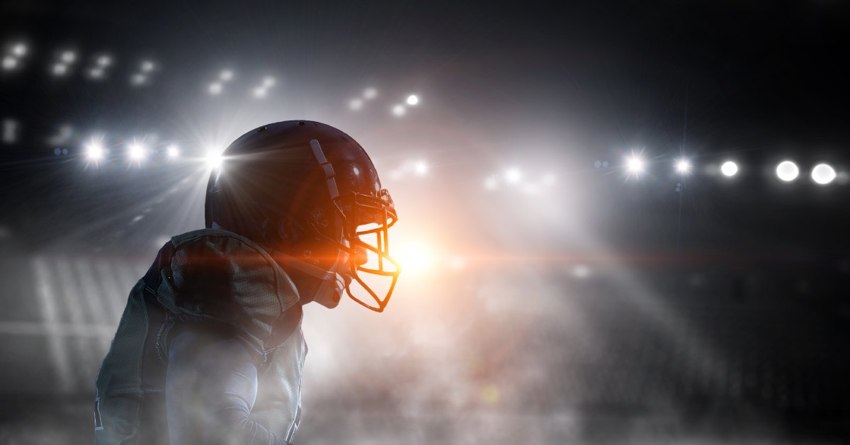 A side view of an American football player with a black helmet and white jersey under the lights of a football stadium.