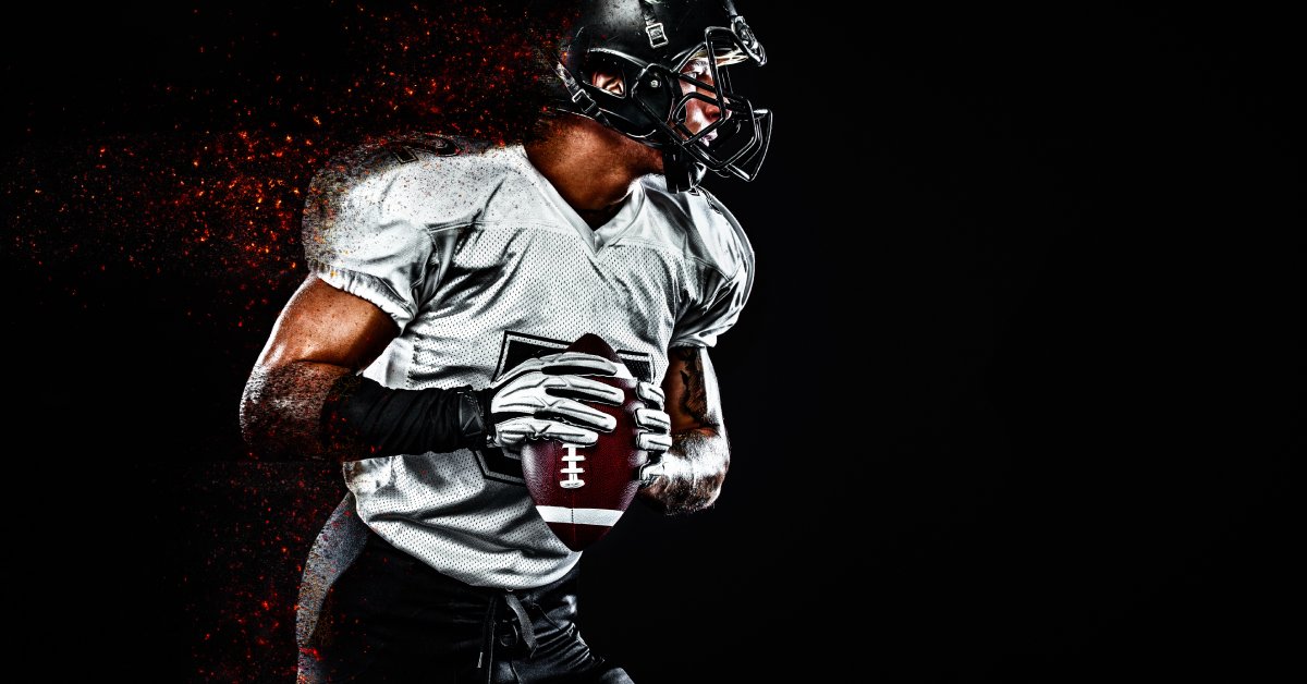 An American football quarterback against a black background, in a white jersey and black helmet, dropping back to pass the ball.