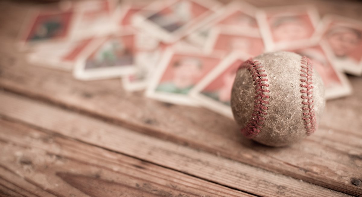 An old, dirty, and used baseball in focus resting on a dirty wooden table with old baseball trading cards blurred behind it.